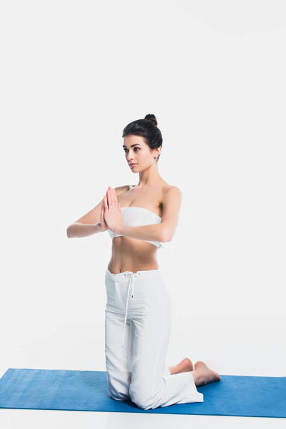 Femme brune avec des mains priantes pratiquant le yoga sur tapis de fitness isolé sur blanc  - Photo, image