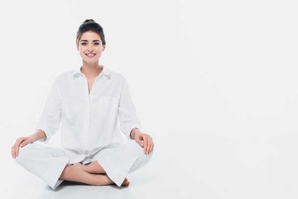 Mujer alegre sentada en postura de yoga y sonriendo a la cámara sobre fondo blanco  - Foto, imagen