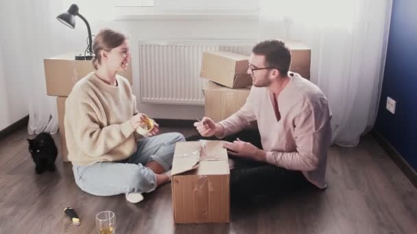 Joyeux couple emballant des boîtes en carton, préparez-vous à déménager, famille assise sur le sol dans le salon le jour du déménagement - Séquence, vidéo