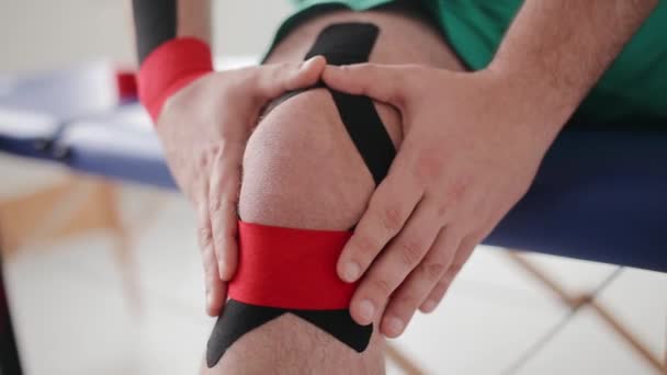 Primer plano del hombre que sufre de dolor en la rodilla herida después de lesiones deportivas. Hombre deportista fija cinta de kinesiología alrededor de la rodilla. Síntomas de dolor: dolor de rodilla. Conceptos de salud y medicina - Metraje, vídeo