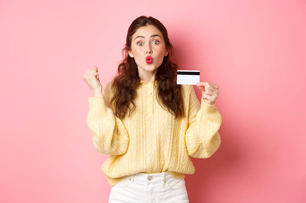 Podekscytowany dziewczyna shopper mówi tak, co pięść pompy i wyglądać zdumiewający, pokazując plastikową kartę kredytową i gotowy do zakupów, stojąc na różowym tle - Zdjęcie, obraz