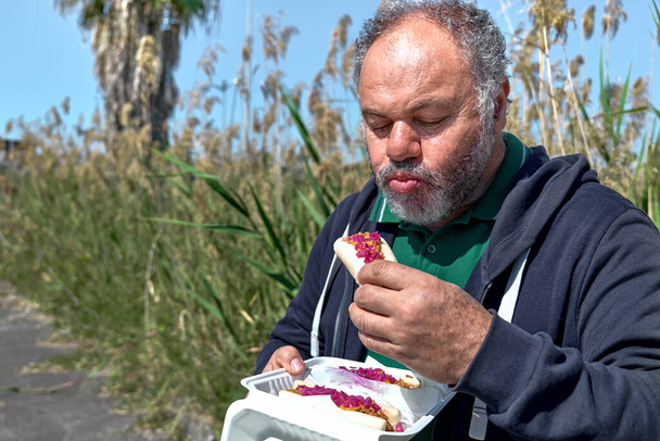 Αστεία γενειοφόρος ώριμος άνθρωπος τρώει vegan χοτ ντογκ στο δρόμο κοντά pampass γρασίδι. Ανθυγιεινό φαγητό. Φάστφουντ. Τροφή για το πακέτο. - Φωτογραφία, εικόνα