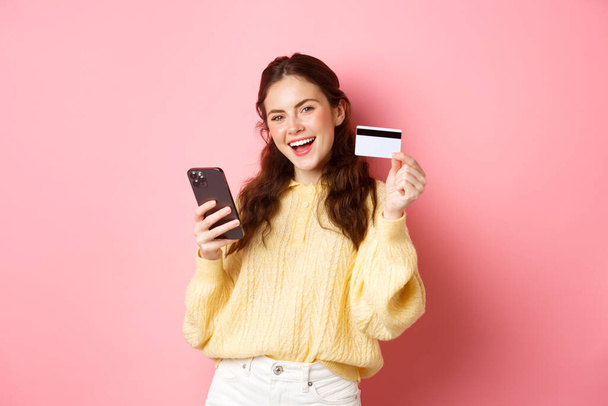 Τεχνολογία και online αγορές. Ευτυχισμένη και ικανοποιημένη πελάτισσα, που δείχνει πλαστική πιστωτική κάρτα και χρησιμοποιεί εφαρμογή κινητού τηλεφώνου για να πληρώσει online, στέκεται απέναντι σε ροζ φόντο - Φωτογραφία, εικόνα