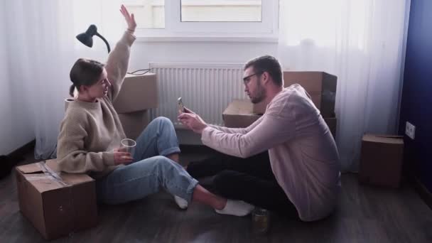 Happy couple relaxes after moving in a new home. Couple sits on floor and celebrates their moving in a new home. Man drinks an alcohol and takes a photo of his wife - Footage, Video