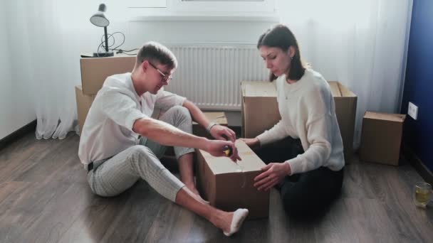 Счастливая пара открывает распакованные картонные коробки после переезда, семья сидит на полу в гостиной в день переезда в новый дом - Кадры, видео