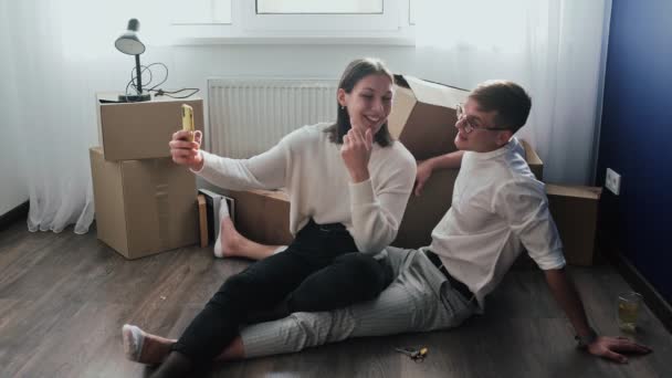 Couple heureux se détend après avoir déménagé dans une nouvelle maison. Couple est assis sur le sol et célèbre leur déménagement dans une nouvelle maison. Ils enregistrent des selfies vidéo au téléphone - Séquence, vidéo