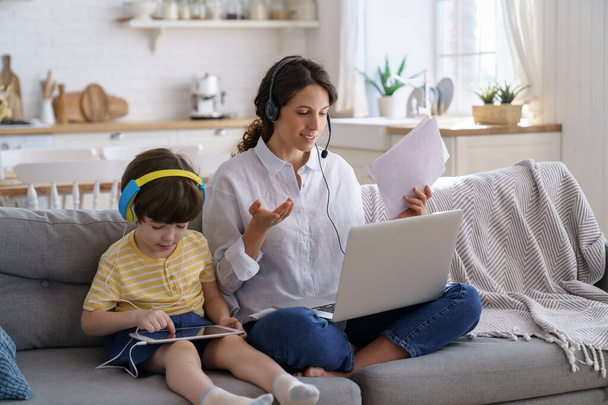 Szabadúszó anya ül kanapén otthon iroda zárlati munka során laptop, gyerek játszik tabletta - Fotó, kép