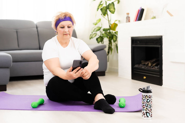 idősebb nő edzőterem otthon gazdaság smartphone. Végzős női edzés fitnesz alkalmazással a telefonban. Fogalom az idősekről és a sportról. - Fotó, kép