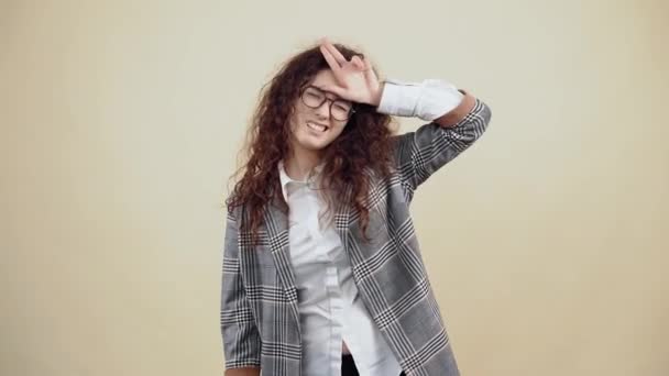The bored, crazy young woman with her fingers emits rhino horns, fooling around. Cretaceous in gray jacket and white shirt, with glasses posing isolated on a beige background in the studio. - Footage, Video