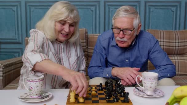 Jubilados pareja de ancianos hablando de beber té, jugar al ajedrez en la sala de estar moderna sala de estar juntos - Imágenes, Vídeo