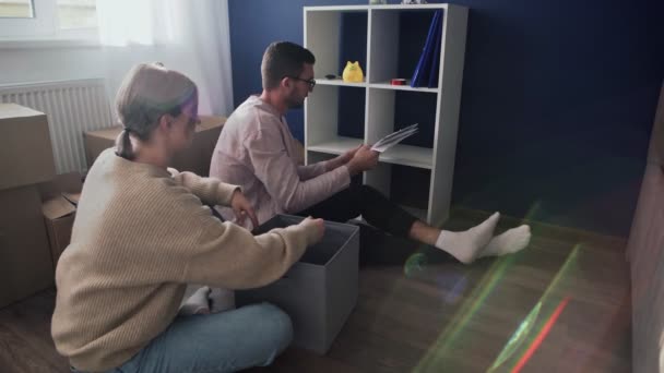 Happy couple open packing cardboard boxes after relocation, family sitting on floor in living room and puts documents from shelf to box - Footage, Video