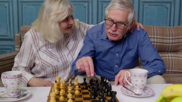 Uśmiechnięta starsza para dziadek babcia spoczywa na kanapie pije kawę, gra w szachy w domu - Materiał filmowy, wideo