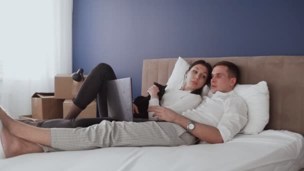 Happy couple relaxes after moving in a new home. Couple lays on bed and looks at notebook screen - Footage, Video