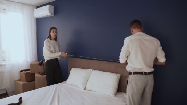 Una joven pareja se prepara para mudarse. Hombre y mujer usan cinta métrica para medir la cama - Metraje, vídeo