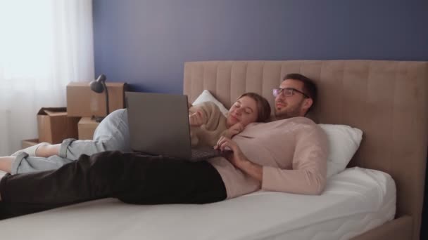 Jong stel ontspant na het verhuizen in een nieuw huis. Paar ligt op bed en kijkt naar notebook scherm - Video