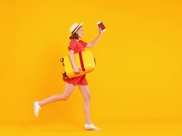Plná délka šťastný legrační mladá žena s lístky a pas s úsměvem při běhu na žlutém pozadí a tahání zavazadla - Fotografie, Obrázek