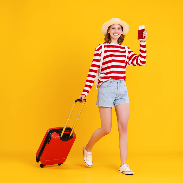 Full length felice giovane donna divertente con biglietti e passaporto sorridente mentre cammina sullo sfondo giallo e tirando bagagli - Foto, immagini