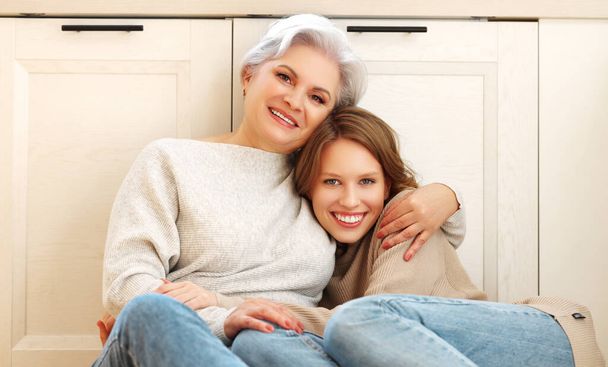 Smiling young woman and gray haired mother in modern casual outfits sitting comfortably in an embraceon floor and looking at camera while spending time together in home kitchen - Photo, Image