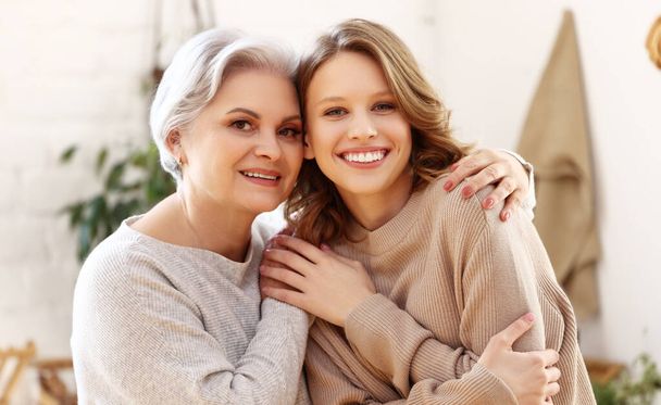 Cheerful young woman hugging aged mother sitting on sofa   while resting together in living room at home - Photo, Image