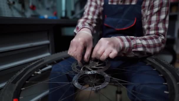 自転車修理店で働く男性整備士、特別なツールを使用して機械修理自転車、保護手袋を身に着けている。若い魅力的なサービスマンは、自分のワークショップでお客様の自転車ホイールを固定 - 映像、動画