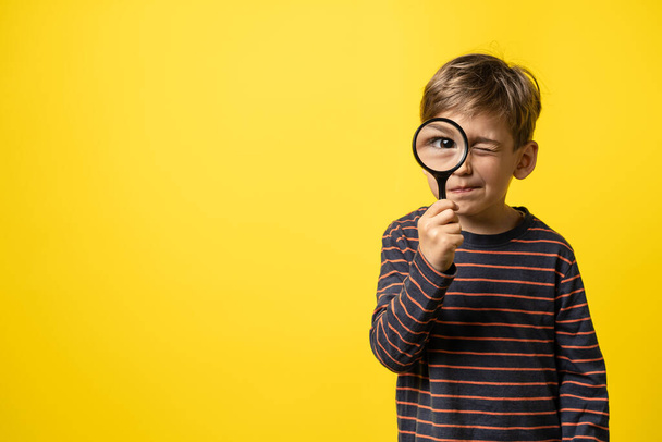 Front view portrait of small caucasian boy curious child holding a magnifying glass for reading in hand inspecting or investigating to have close look with eye in focus - copy space yellow background - Фото, изображение