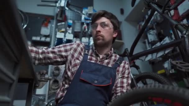 Młody biały mechanik naprawiający koło rowerowe w warsztacie rowerowym. Naprawiam rower. Troszczę się o ciebie. mechanik pracujący w garażu. Pracownicy naprawiają koła zębate. Temat małych przedsiębiorstw - Materiał filmowy, wideo