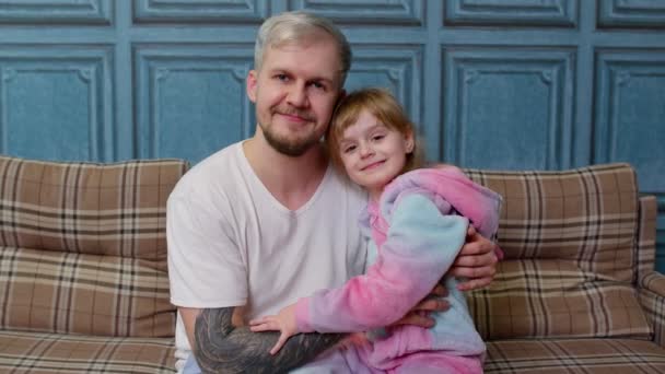 Отец и маленькая дочь дочь ребенок в пижаме сидеть на диване в комнате улыбаясь, глядя в камеру - Кадры, видео