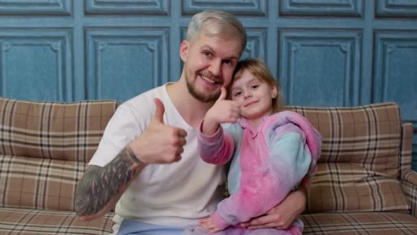 Padre e hija pequeña niño en pijama sentarse en el sofá en la habitación sonriendo, mostrando los pulgares hacia arriba - Imágenes, Vídeo