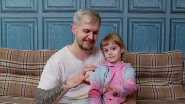 パジャマの父と子の娘子供はソファの上に笑顔で座って胸近くの形の心のサインを作る - 映像、動画