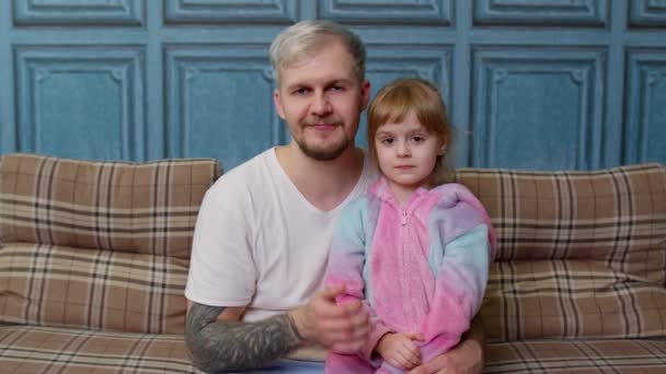 Πατέρας και παιδί κορίτσι κόρη με πιτζάμες κάθονται στον καναπέ στο δωμάτιο χαμογελώντας, φυσώντας στέλνοντας ένα φιλί αέρα - Πλάνα, βίντεο