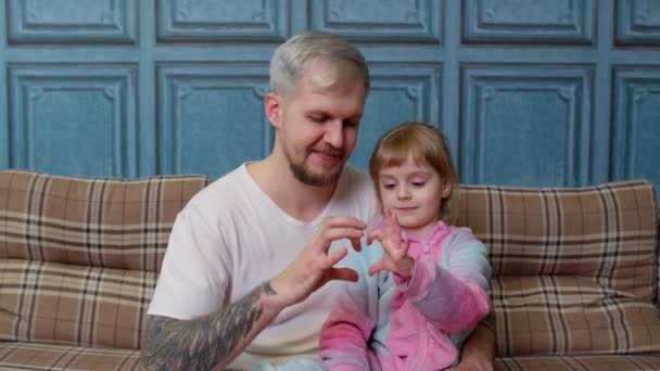 Ojciec i dziecko córka dziecko w piżamie siedzieć na kanapie uśmiechając się, co znak kształtu serca w pobliżu klatki piersiowej - Materiał filmowy, wideo