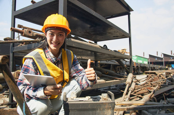 Egy ázsiai női gyári munkás kemény kalapban és sárga mellényben ül egy táblával a kezében, és egy használt ipari elemet vizsgál egy halom régi anyaggal. Ipari hulladékgazdálkodás. - Fotó, kép