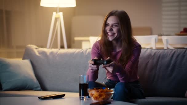 Jonge emotionele dame spelen videospelletjes op gamepad thuis, genieten van vrijdagavond met snacks, slow motion - Video