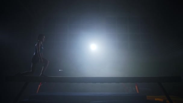 deportista profesional está realizando en la viga de equilibrio, chica joven está caminando sobre aparato de gimnasia artística en salón oscuro - Imágenes, Vídeo