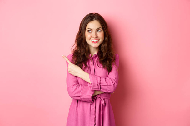 Όμορφη κοπέλα αίγλη με μακιγιάζ, φορώντας στολή άνοιξη, δείχνοντας και κοιτάζοντας κατά μέρος το λογότυπο, δείχνει πανό στο αριστερό copyspace, στέκεται πάνω σε ροζ φόντο - Φωτογραφία, εικόνα