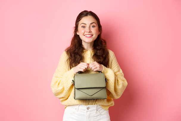 Beautiful lady holding her purse in hands near chest, smiling and looking excited at camera, waiting for something with temptation, standing against pink background - Photo, Image