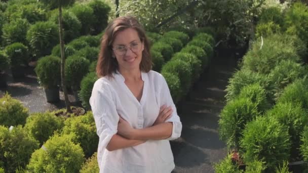 Medium PAN portrait with slowmo of smiling caucasian woman in eyeglasses standing with hands folded in beautiful green garden looking at camera - Footage, Video