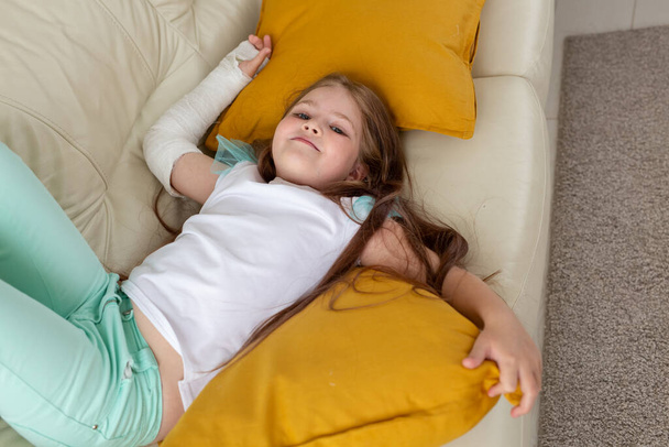 Gyermek gipsszel a törött csuklóján vagy karján mosolyog és szórakozik a kanapén. Pozitív hozzáállás, felépülés és gyerekkoncepció. - Fotó, kép