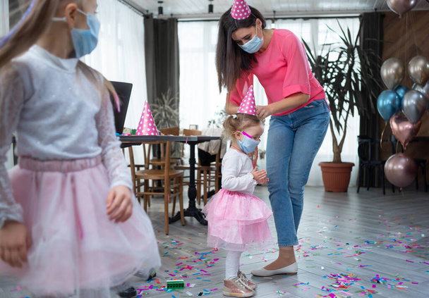 Κατά τη διάρκεια του πάρτι, η μητέρα ντύνει το κορίτσι με μια ιατρική μάσκα. Έννοια εορτασμού γενεθλίων. Αντιγραφή χώρου. - Φωτογραφία, εικόνα