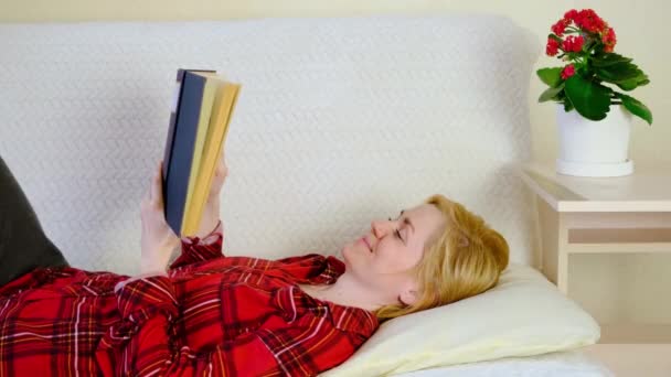 Щаслива молода кавказька жінка в червоній сорочці сидить на легкому дивані з книжкою. Поняття читання, літератури, відпочинку вдома.. - Кадри, відео
