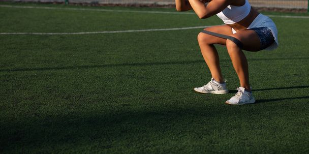 Muskularna, wysportowana kobieta robi przysiady z opaską fitness na boisku. Puste miejsce  - Zdjęcie, obraz