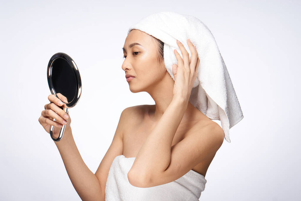 веселая красивая женщина смотрит в зеркало и полотенце на голове уход за кожей спа-процедуры легкий фон - Фото, изображение