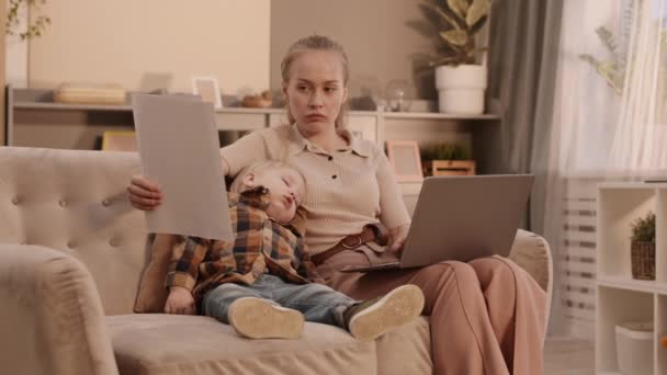 Μέτριο πλάνο της νεαρής Καυκάσιας γυναίκας που κάθεται στον καναπέ στο σαλόνι, χρησιμοποιώντας φορητό υπολογιστή, κοιτάζοντας τα χαρτιά της επιχείρησης, μιλώντας στο κινητό τηλέφωνο, ο μικρός γιος της κοιμάται κοντά - Πλάνα, βίντεο
