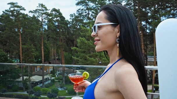 боковой вид счастливой брюнетки в солнечных очках, держащей стакан с коктейлем - Фото, изображение