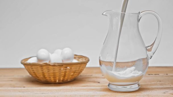 milk pouring into glass jar near wicker basket with white eggs on wooden table isolated on grey - Photo, Image