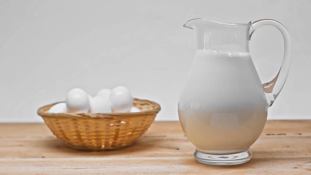 стеклянная банка со свежим молоком возле плетеной корзины с белыми яйцами на деревянном столе, изолированном на сером - Фото, изображение