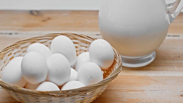 木のテーブルの上に新鮮な牛乳とガラス瓶の近くのウィッカーバスケットに鶏の卵の山 - 写真・画像