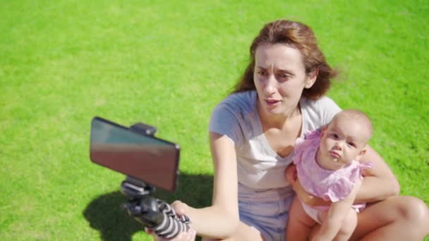 Mother blogger with little daughter having fun records a video blog on green grass. - Footage, Video