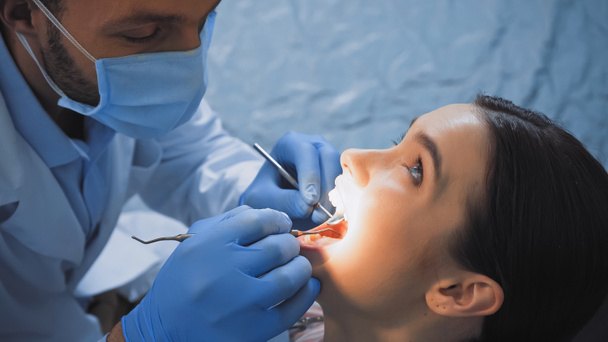 estomatólogo en máscara médica examinar los dientes del paciente con instrumentos dentales - Foto, Imagen