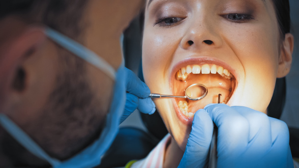 стоматолог в медицинской маске, изучающий зубы женщины, размытый передний план - Фото, изображение